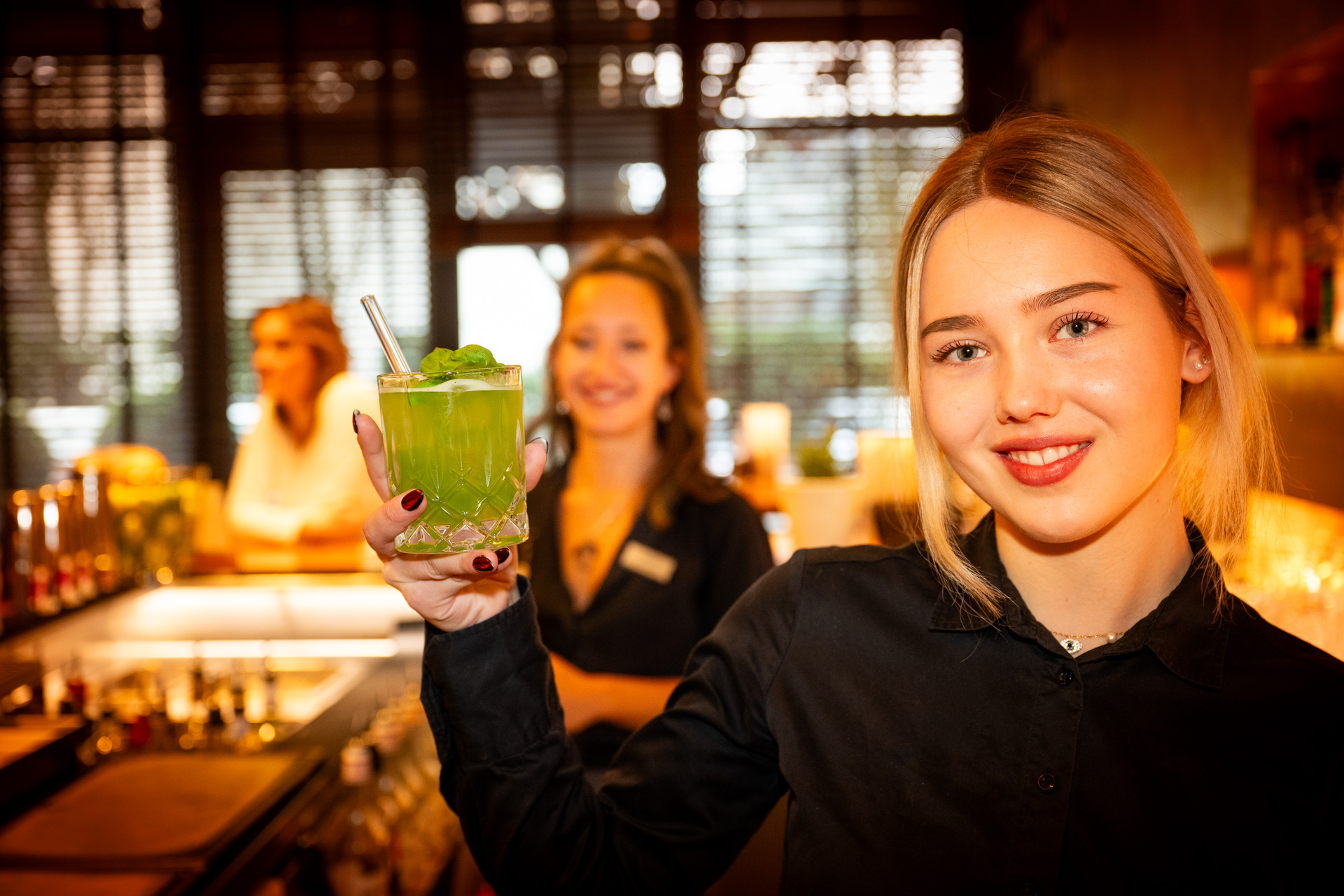 Barkeeperin hält lächelnd einen grünen Cocktail. Eine weitere Barkeeperin im Hintergrund.