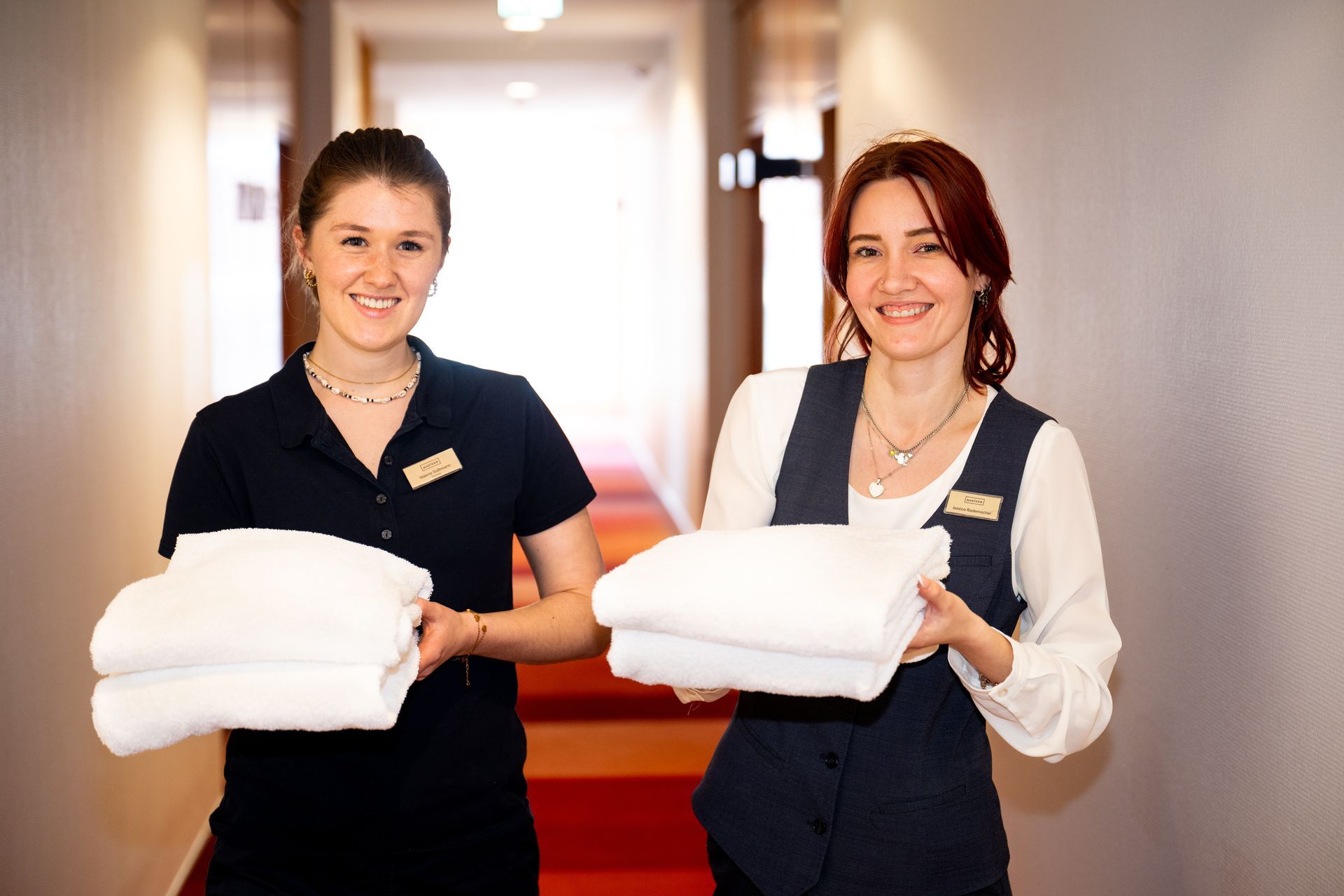 Two maids are carrying stacks of towels in a hotel hallway.