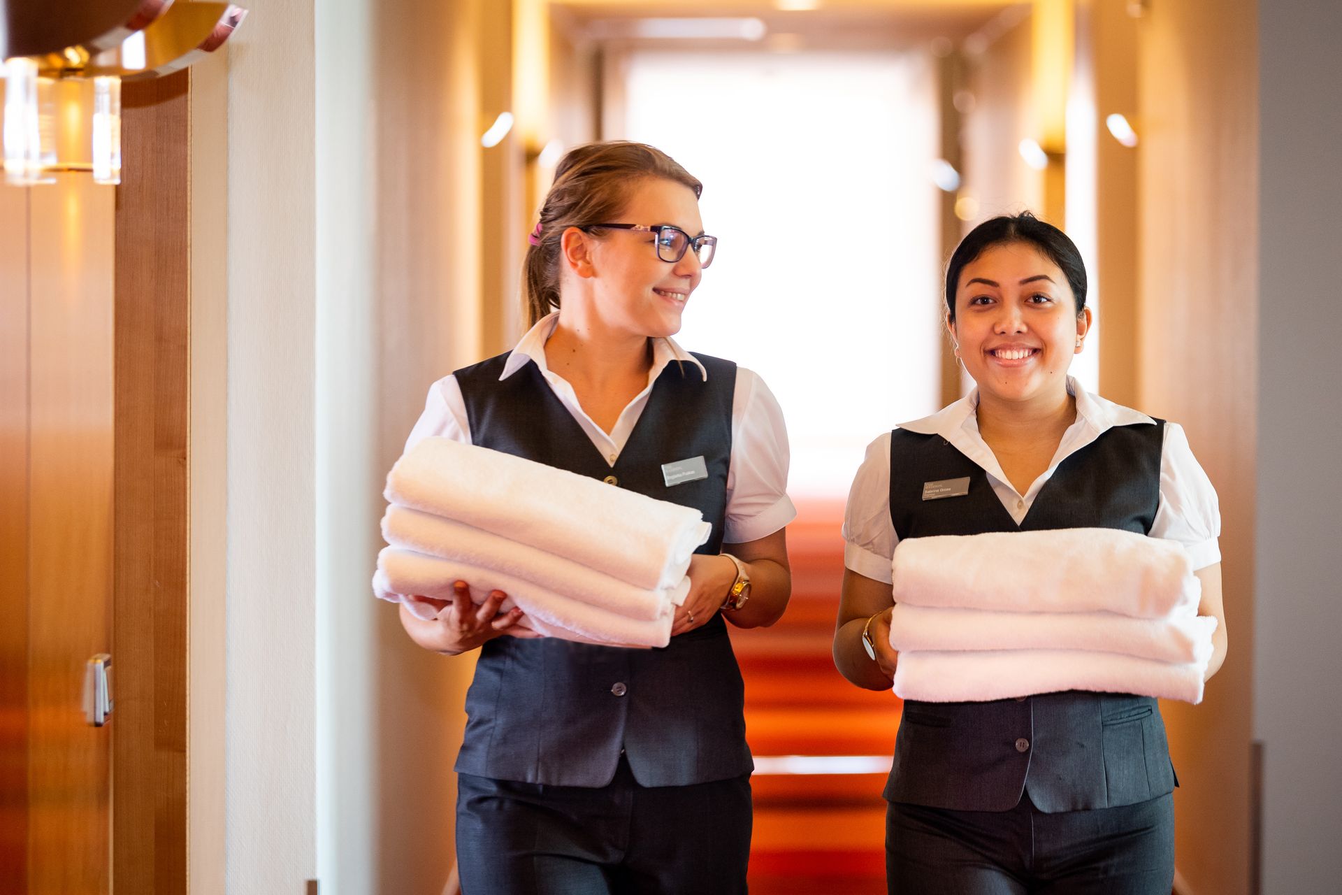 Two maids are carrying stacks of towels in a hotel hallway.