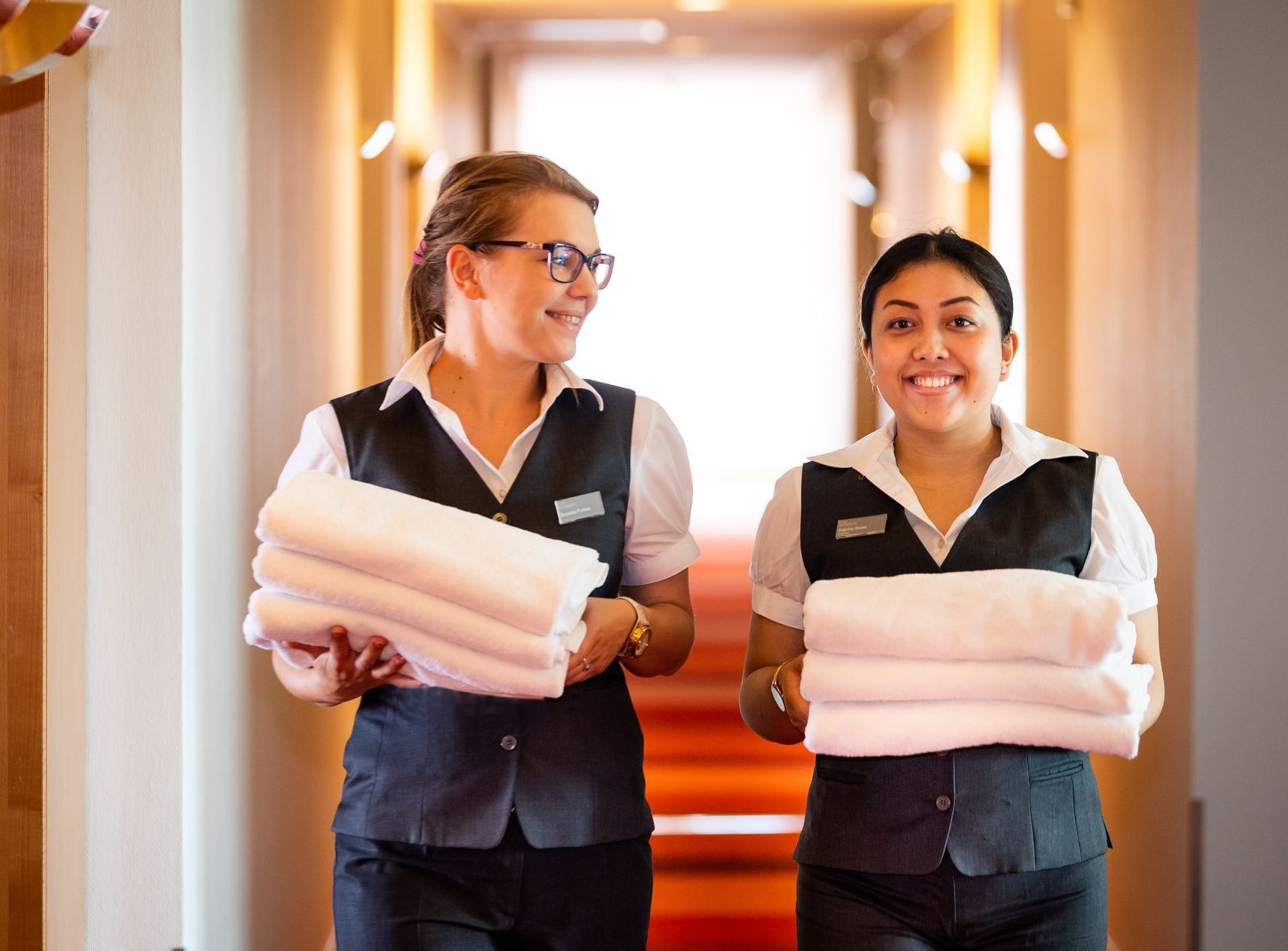 Zwei Zimmermädchen tragen Stapel von Handtüchern durch einen Hotelflur.