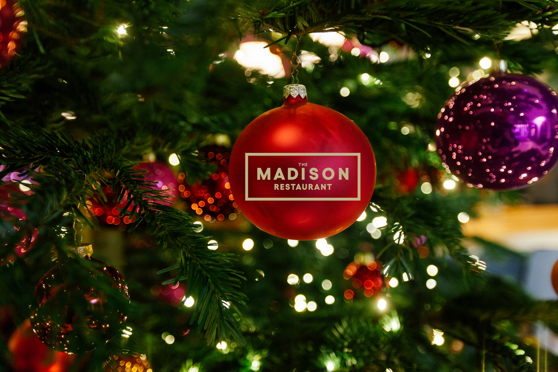 Ein roter Christbaumschmuck mit dem Logo von „The Madison Restaurant“, umgeben von Lichtern und anderem Schmuck.