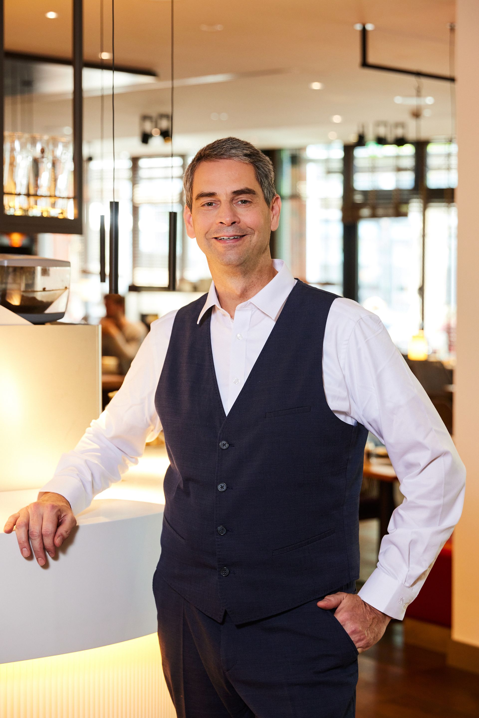 Ein Mann in Weste und weißem Hemd lehnt an einer Theke in einem Restaurant.