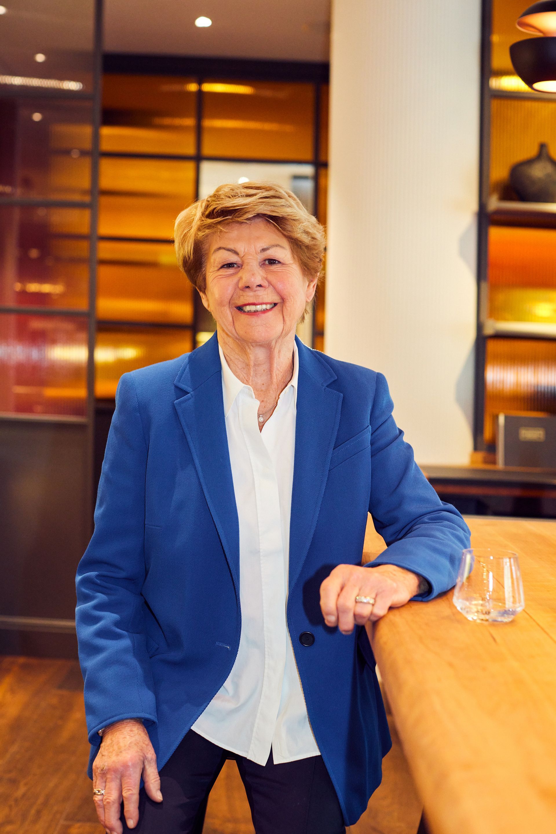A woman is wearing a white shirt and a blazer and leaning against a wooden table in a hotel near Hamburg harbour.
