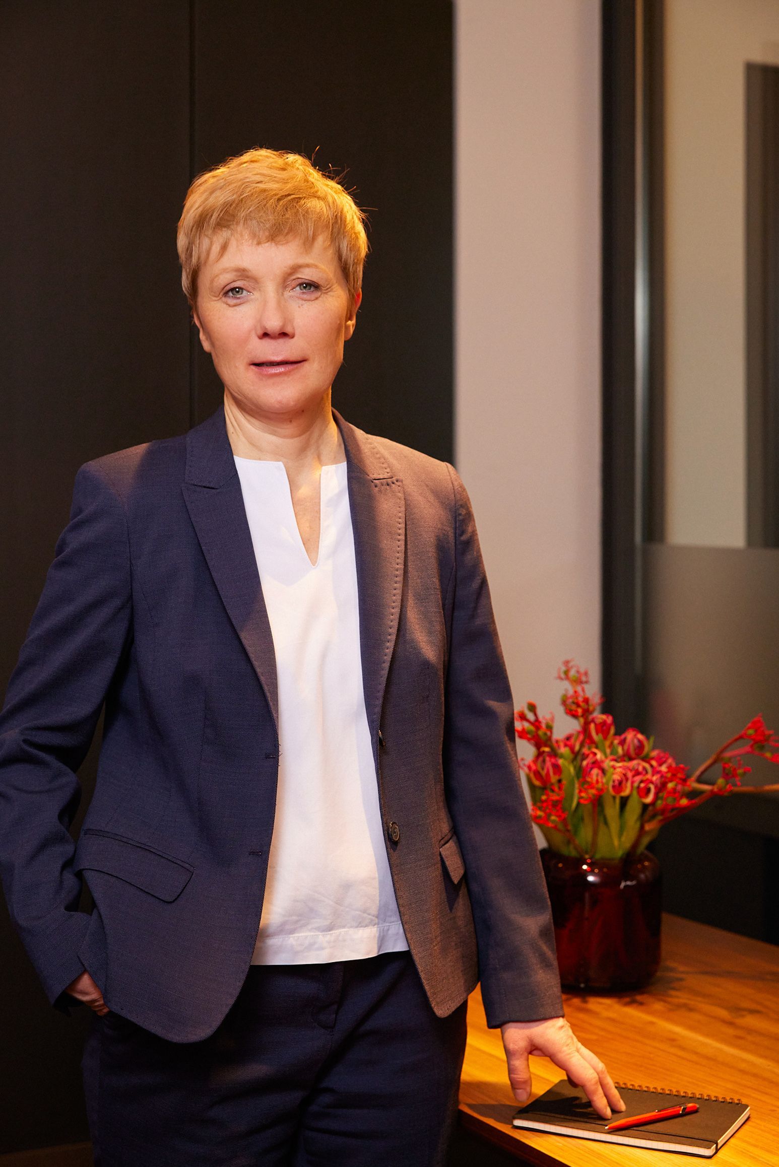 Eine Frau im Anzug steht vor einem Tisch mit Blumen in einer Vase.