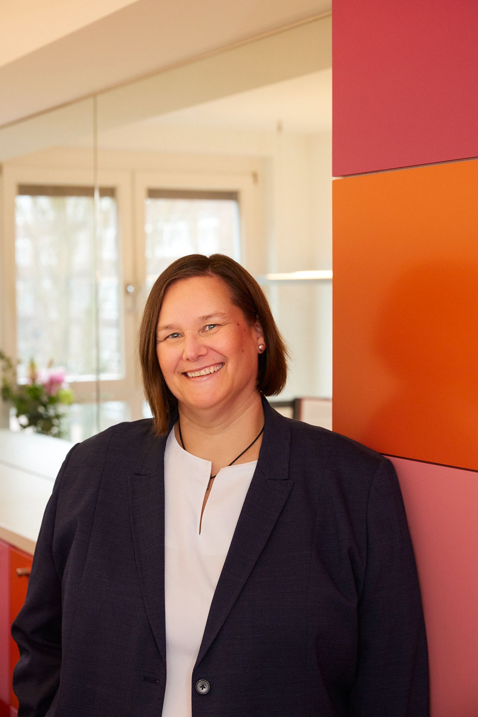 Eine Frau im Anzug lehnt an einer Wand und lächelt.