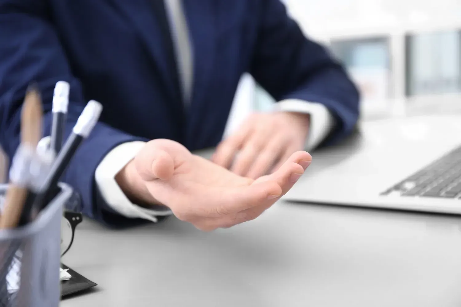 Manos escribiendo con un bolígrafo sobre un documento; al fondo hay una pila de papeles y un teclado.