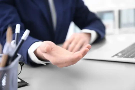 Manos escribiendo con un bolígrafo sobre un documento; al fondo hay una pila de papeles y un teclado.