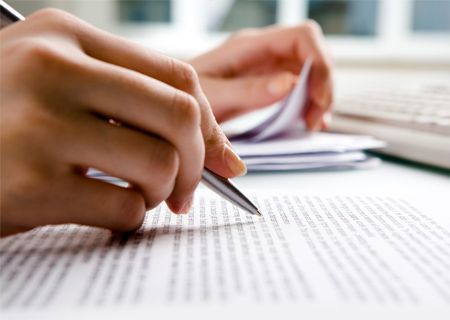 Manos escribiendo con un bolígrafo sobre un documento; al fondo hay una pila de papeles y un teclado.