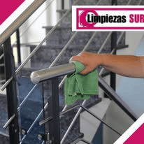 Una mano limpia la barandilla de acero inoxidable de una escalera con un paño verde. En una esquina se ve el logotipo de