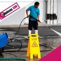 Una persona con una camisa azul utiliza una hidrolavadora para limpiar una superficie de concreto junto a una señal de precaución amarilla.