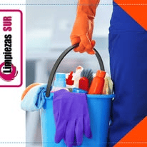 Una persona vestida con un uniforme azul y un guante naranja lleva un cubo azul lleno de diversos productos de limpieza y un guante morado.