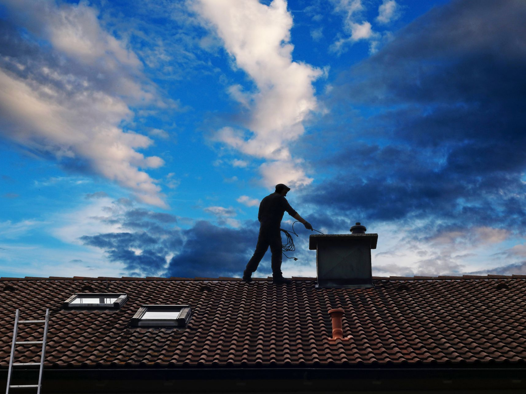 Vers la page Entretien de cheminée. Un homme nettoie une cheminée sur un toit.