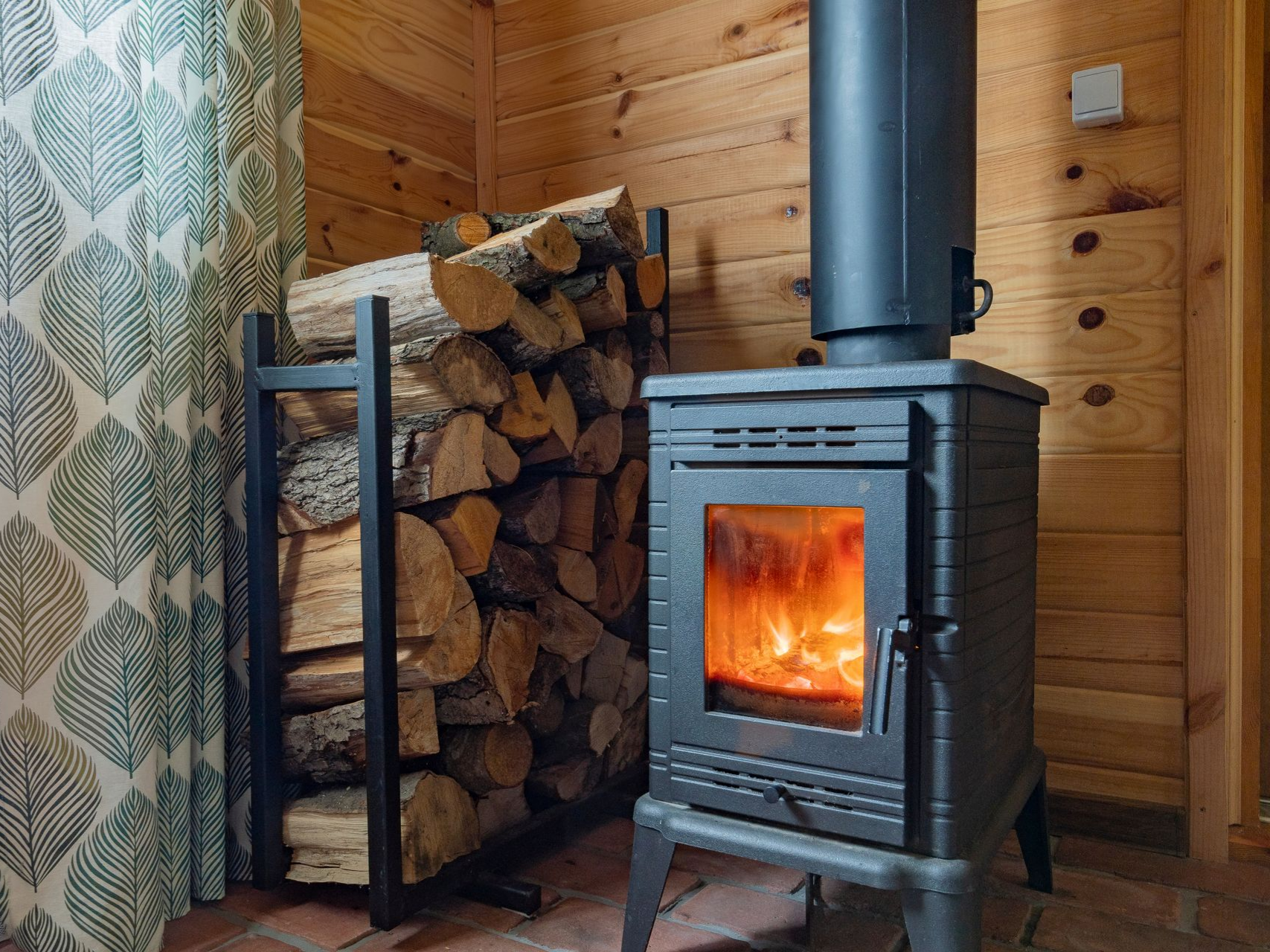 Vers la page Installation de poèle. Un poêle à bois avec des flammes, à côté d'une pile de bois de chauffage.