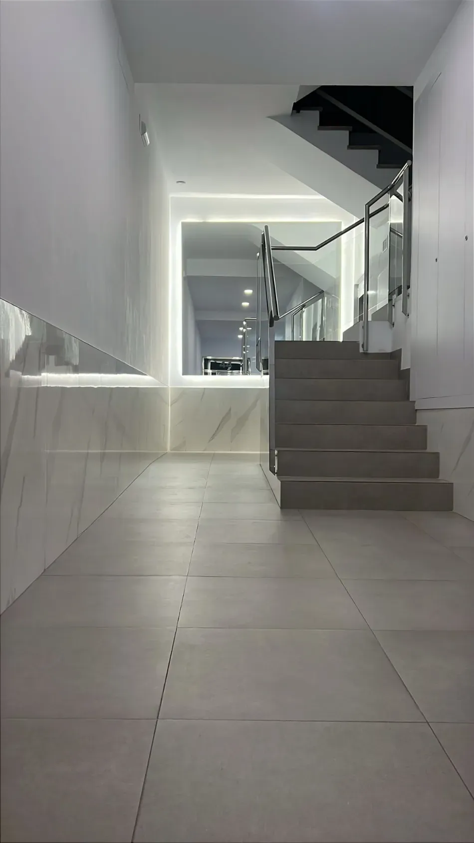 Escalera interior moderna con paredes blancas, escalones grises y un pasillo alicatado que conduce a un rellano superior.