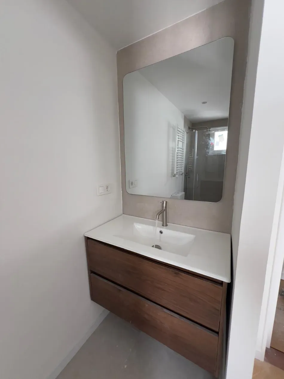 Tocador de baño moderno con lavabo blanco, cajones de madera y espejo grande en una habitación estrecha.