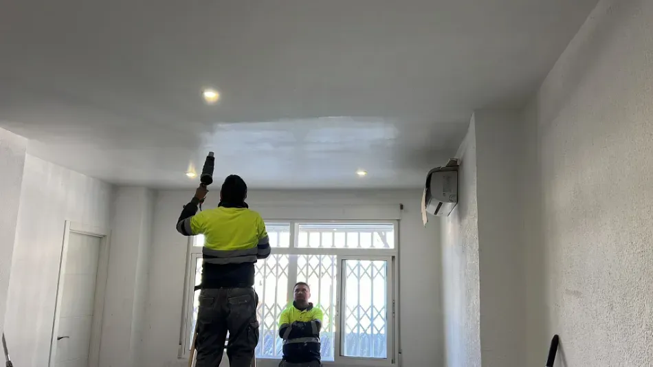 Dos trabajadores con chalecos reflectantes pintan una habitación blanca con rodillo y brocha.