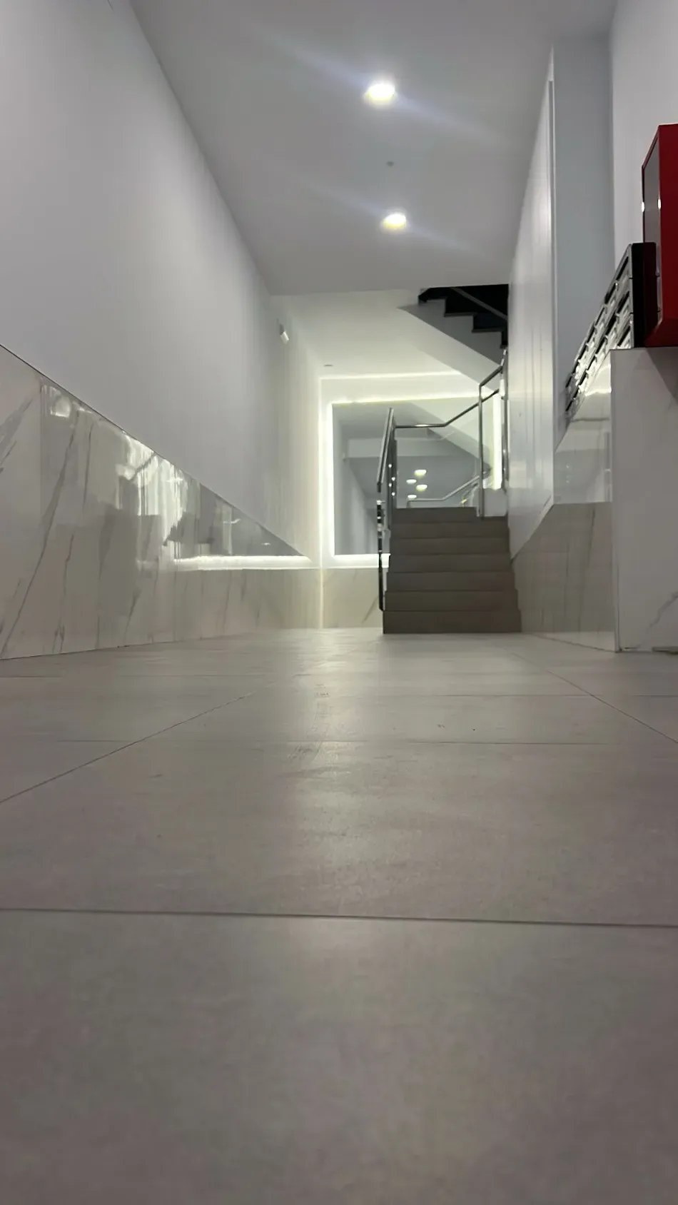 Pasillo moderno con paredes blancas, suelo de baldosas, escaleras y luces de techo brillantes que conducen a un rellano superior.
