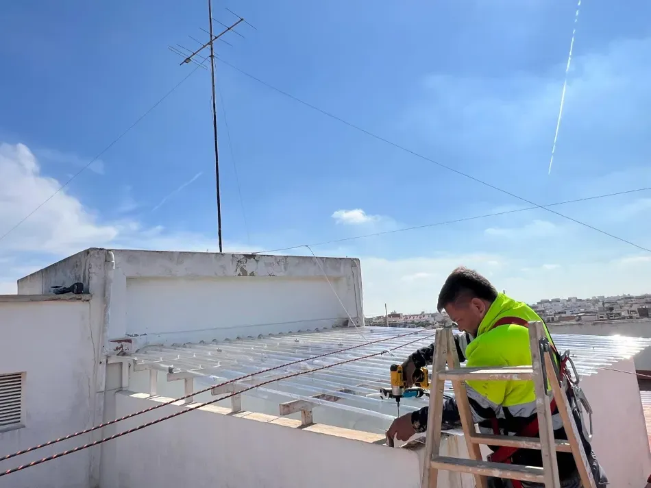 Trabajador con chaleco reflectante reparando equipos en la azotea bajo un cielo azul.