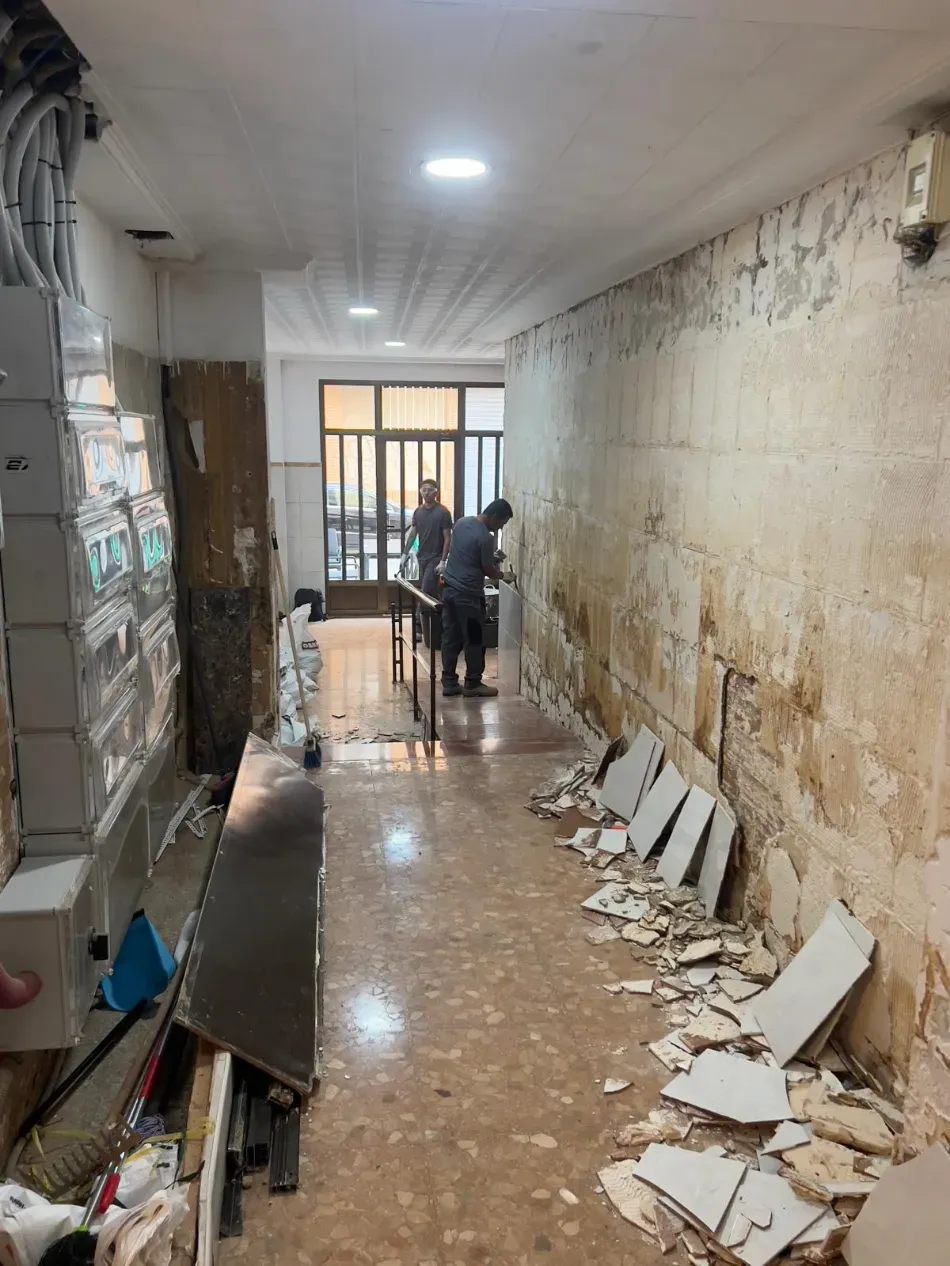 Pasillo estrecho y deteriorado, con paredes desconchadas, escombros esparcidos y dos personas cerca de una mesa al fondo.