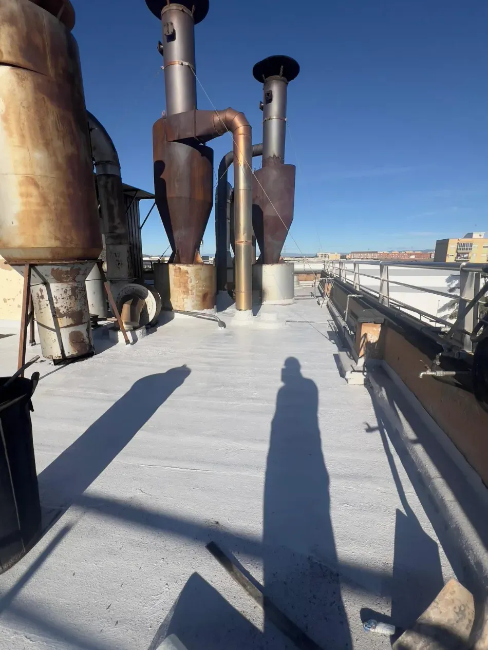 Pasarela en la azotea, junto a conductos de ventilación y tuberías industriales, bajo un cielo azul despejado, con largas sombras en el suelo.