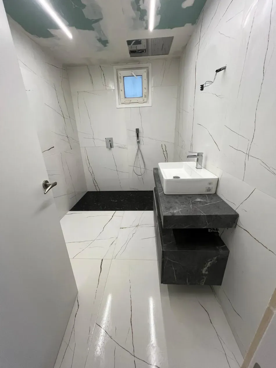 Baño moderno con paredes de mármol blanco, lavabo negro y una pequeña ventana en la parte trasera.