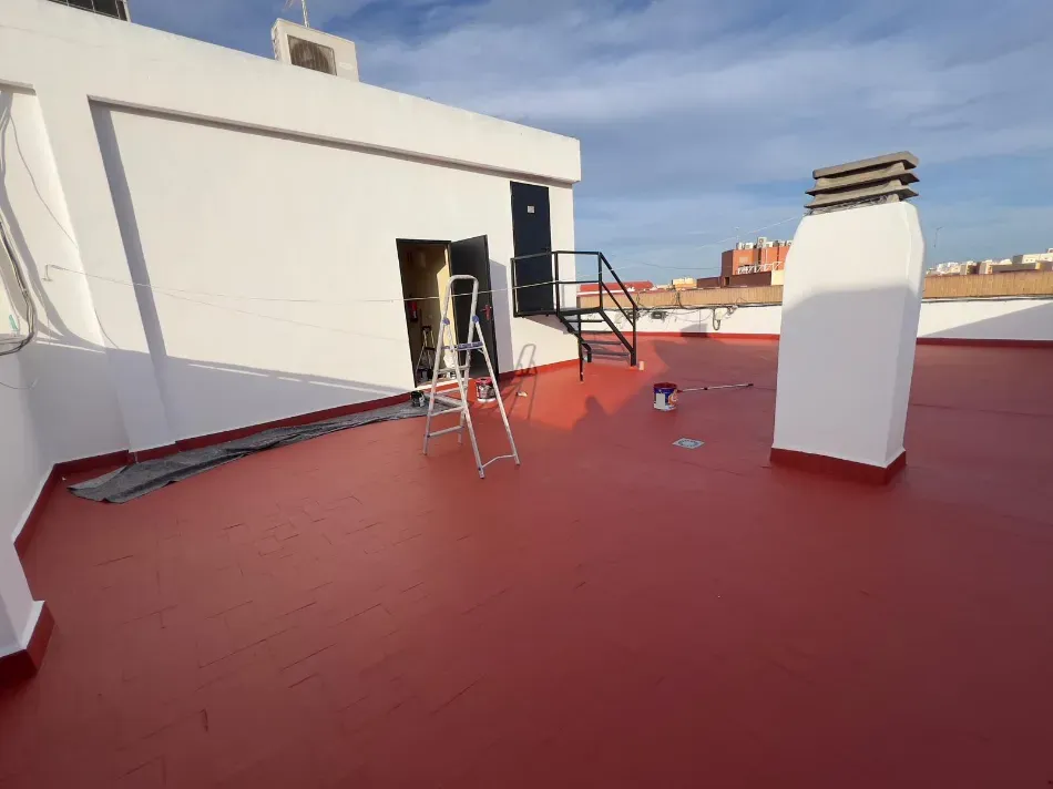 Terraza en la azotea con suelo rojo, paredes blancas, una silla cerca de una puerta abierta y una pequeña chimenea.