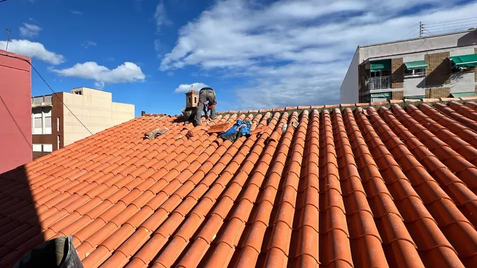 Trabajadores en un tejado de tejas rojas bajo un cielo azul, con sus herramientas y un edificio vecino cerca.