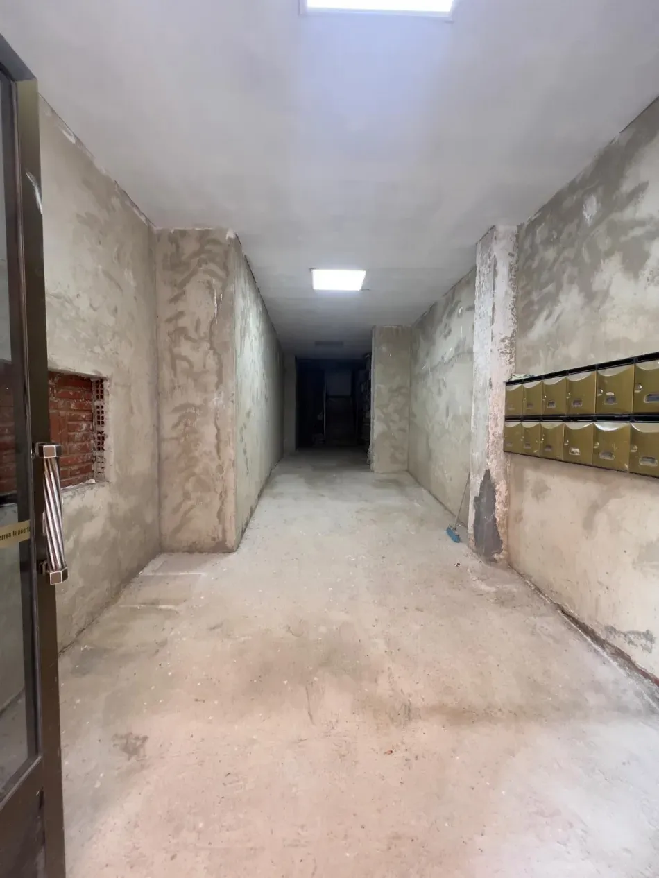 Pasillo estrecho y oscuro con paredes de color beige desgastado, buzones a la derecha y una puerta oscura al final.