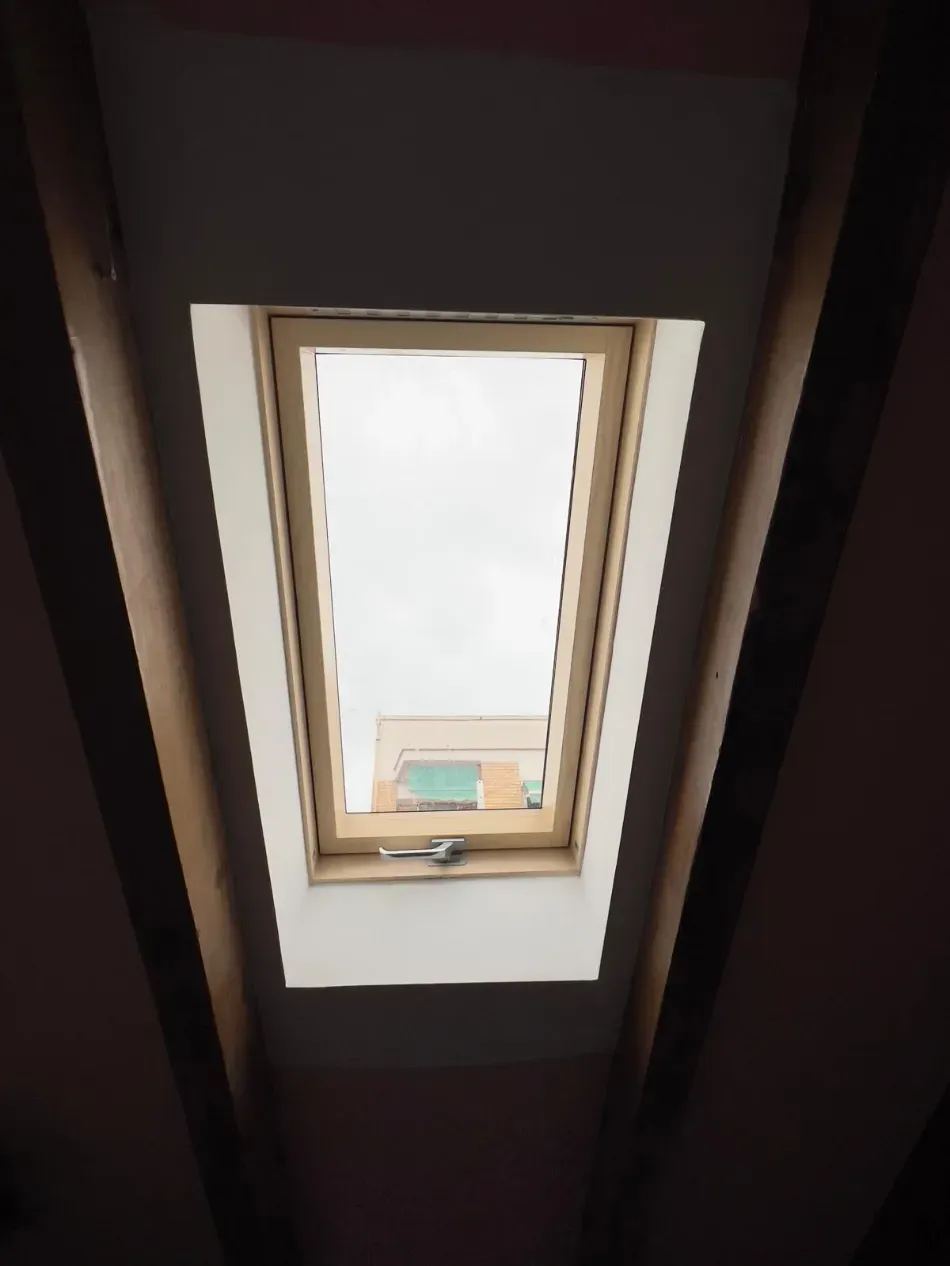 Ventana de techo acristalada en un techo oscuro, que deja ver un cielo nublado y un pequeño edificio debajo.