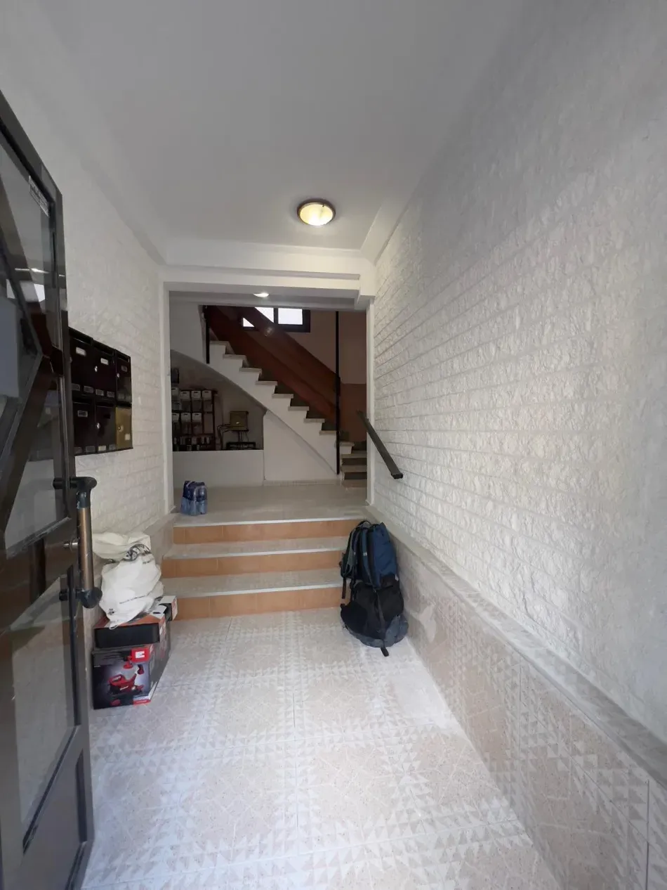 Pasillo estrecho con suelo de baldosas, escaleras al final y una mochila apoyada contra la pared.