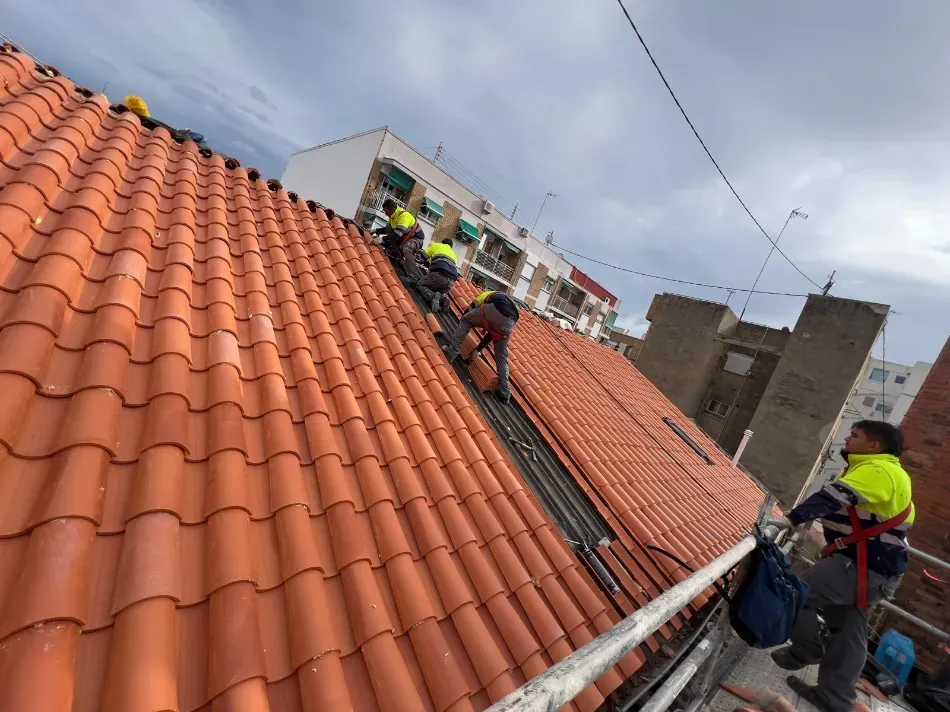 Trabajadores instalando paneles solares en un tejado de tejas rojas con herramientas y equipo de seguridad.