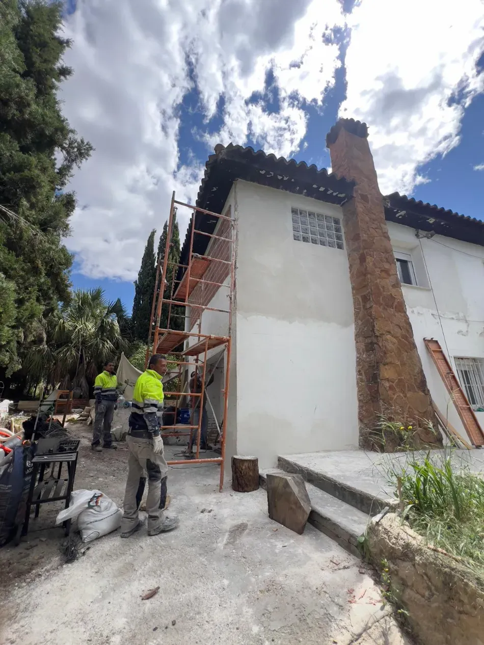 Trabajadores en andamios pintando el exterior de una casa blanca junto a una chimenea de ladrillo bajo un cielo nublado.