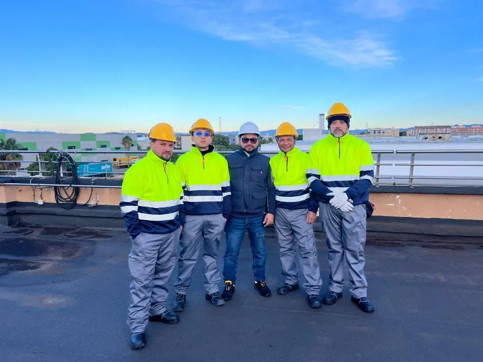 Cinco trabajadores con cascos y chalecos reflectantes están de pie en una azotea con el perfil urbano de la ciudad de fondo.