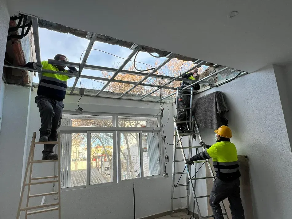 Trabajadores con chalecos reflectantes reparan un techo dañado en una habitación luminosa.