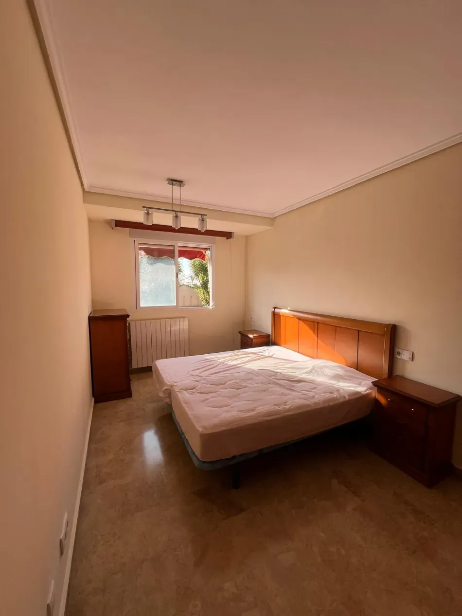 Habitación pequeña sin amueblar con cama, mesitas de noche, cómoda y ventana con radiador.