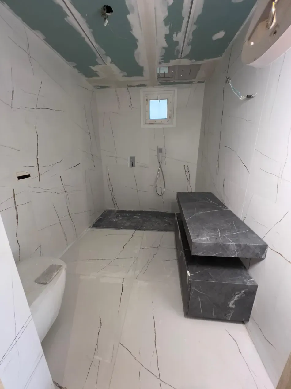 Baño estrecho y sin terminar, con paredes alicatadas en blanco, banco oscuro y una pequeña ventana al fondo.