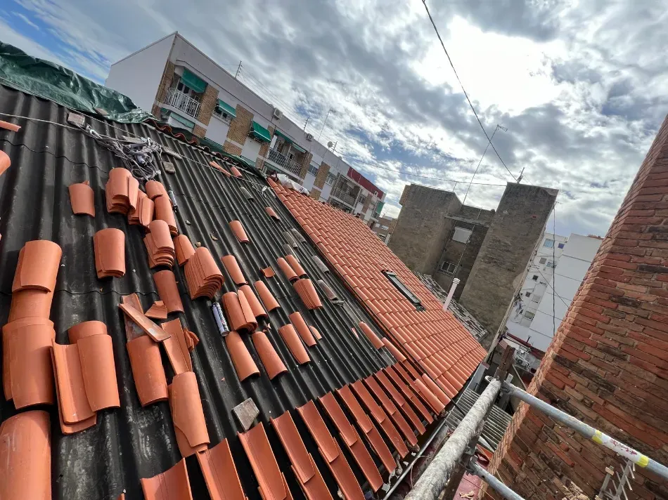 Tejas de arcilla roja esparcidas sobre un tejado empinado bajo un cielo nublado, con edificios cercanos a la vista.