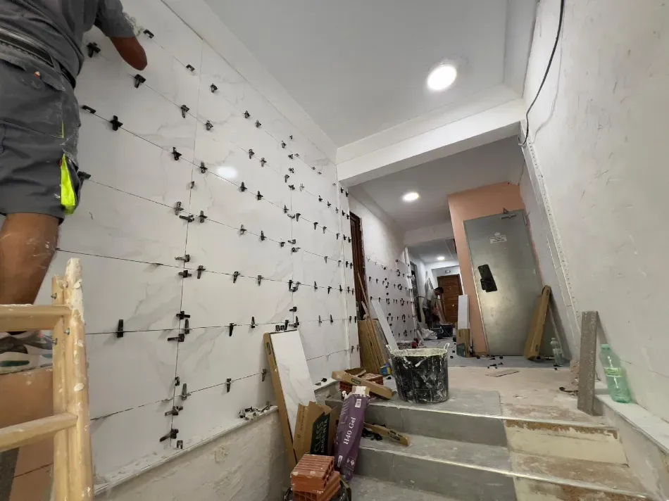 Pasillo en obras: se han retirado los azulejos de la pared, se ven herramientas de construcción y un trabajador parcialmente visible en una escalera.