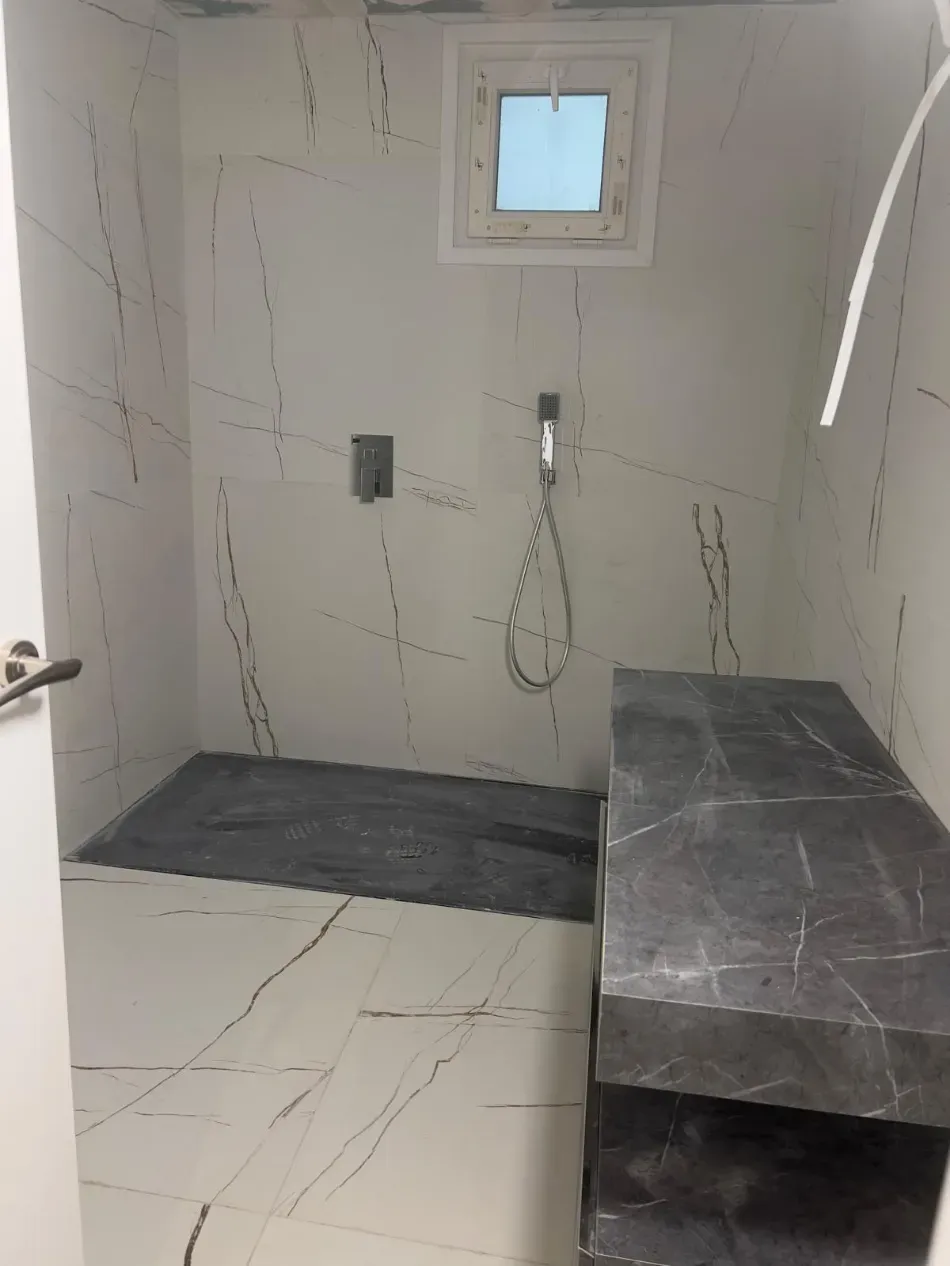 Baño pequeño y moderno con paredes que imitan mármol blanco, una ventana, grifería de ducha y un banco oscuro.