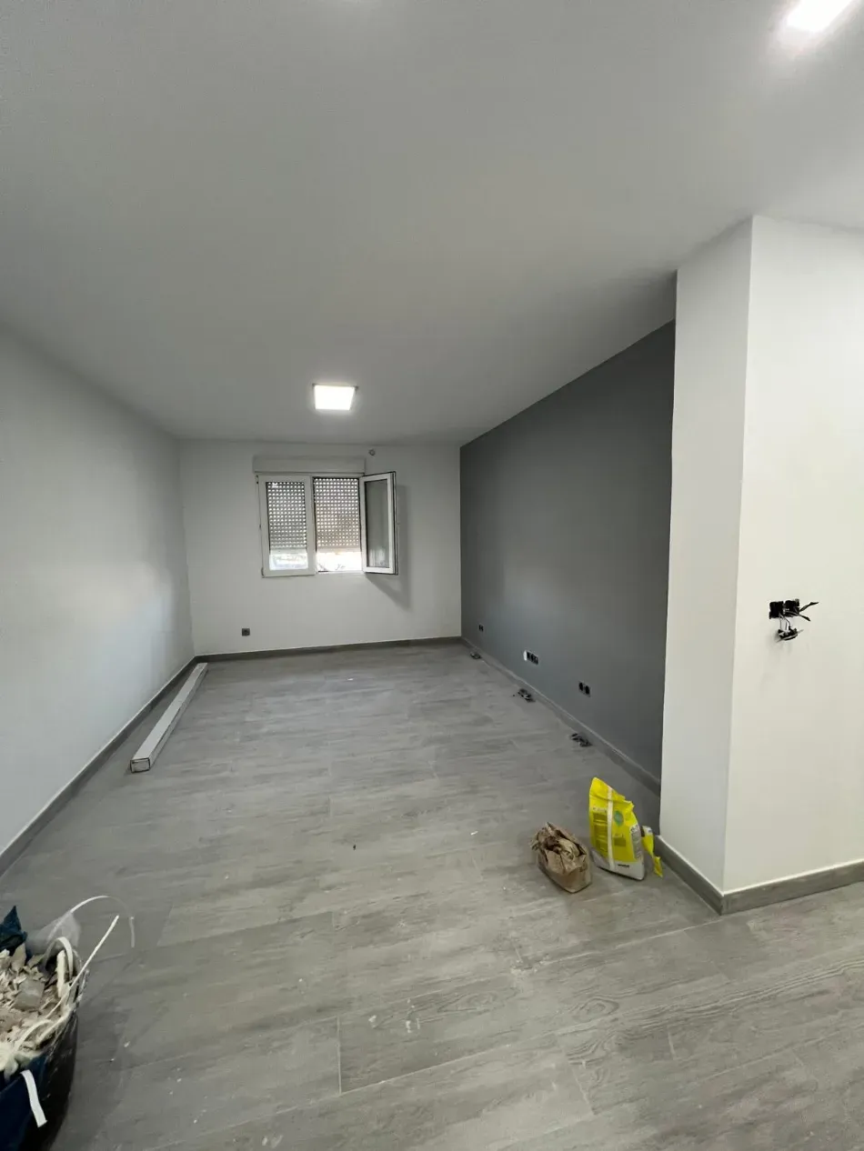Habitación vacía sin amueblar con suelo gris, paredes blancas, una ventana y materiales de construcción cerca de la esquina.