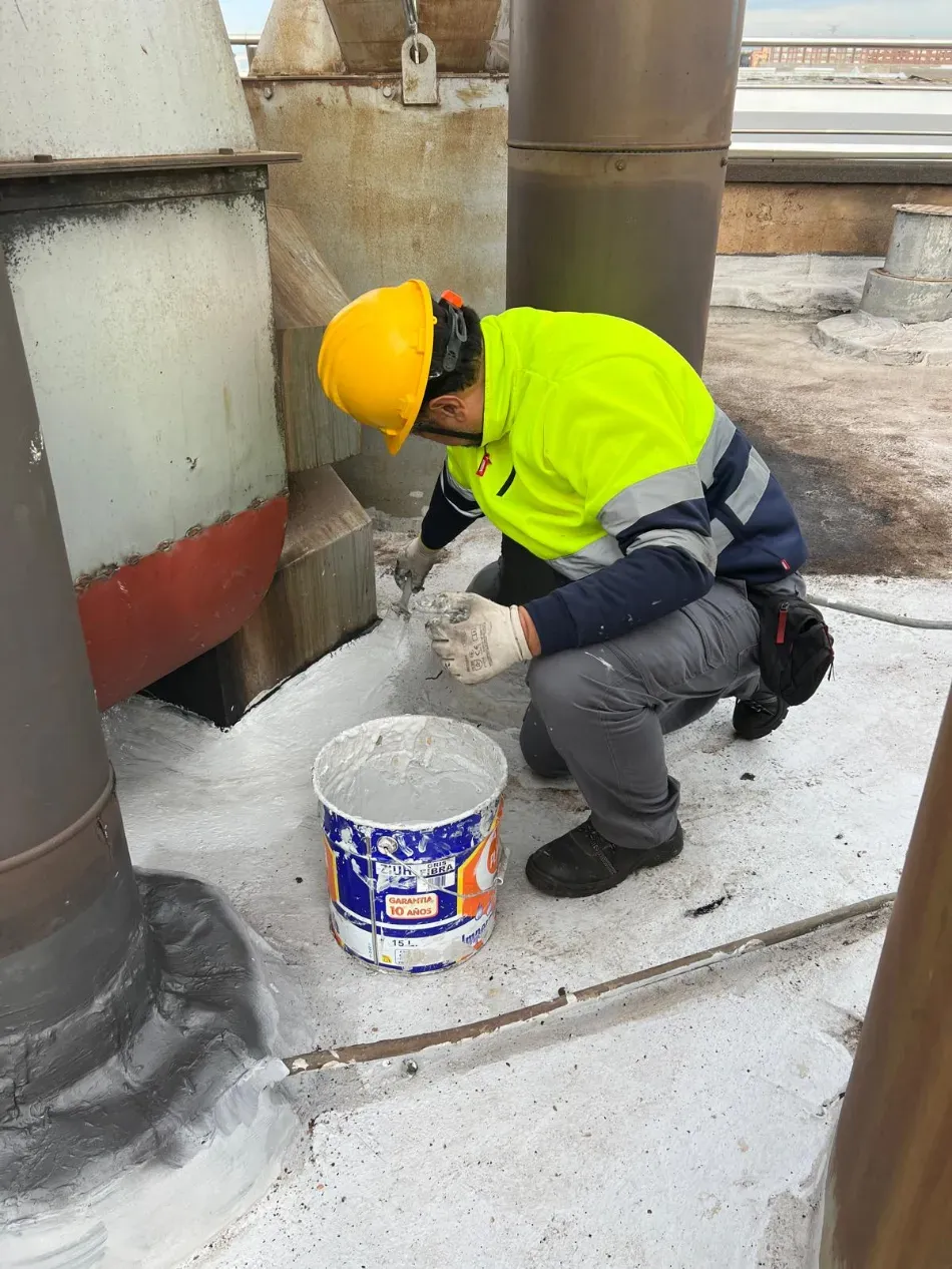 Un trabajador con casco amarillo, arrodillado junto a unas tuberías, aplica una capa de pintura blanca en una zona industrial.