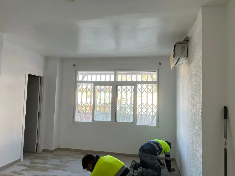Trabajadores instalando el suelo en una habitación luminosa y vacía con paredes blancas y una gran ventana.