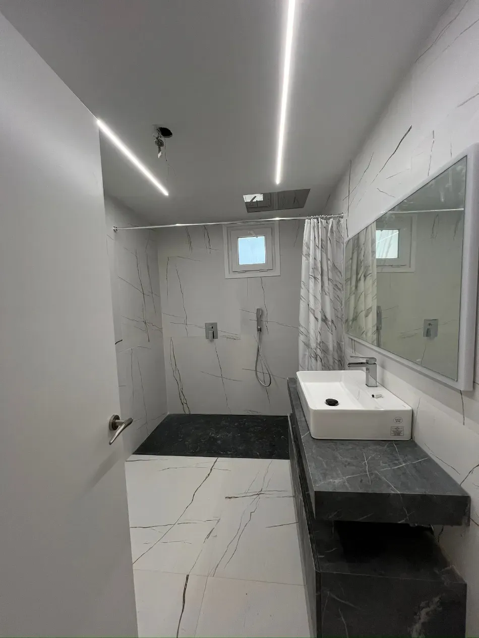 Baño pequeño y moderno con azulejos de mármol blanco, ducha, lavabo, espejo y encimera oscura.