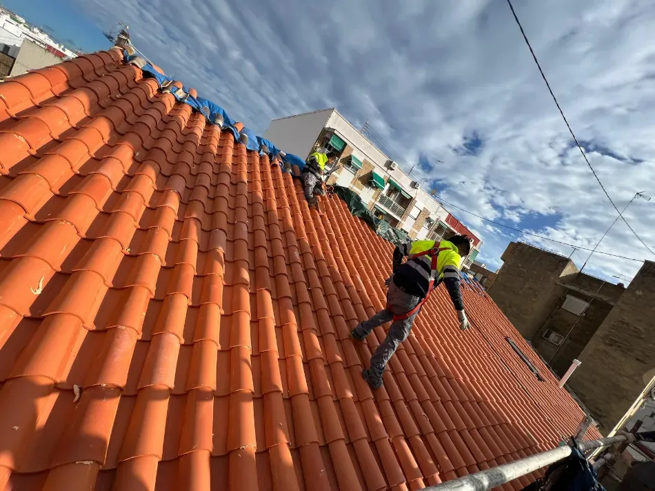 Trabajadores en un tejado de tejas rojas, sujetos con arneses, bajo un cielo nublado.
