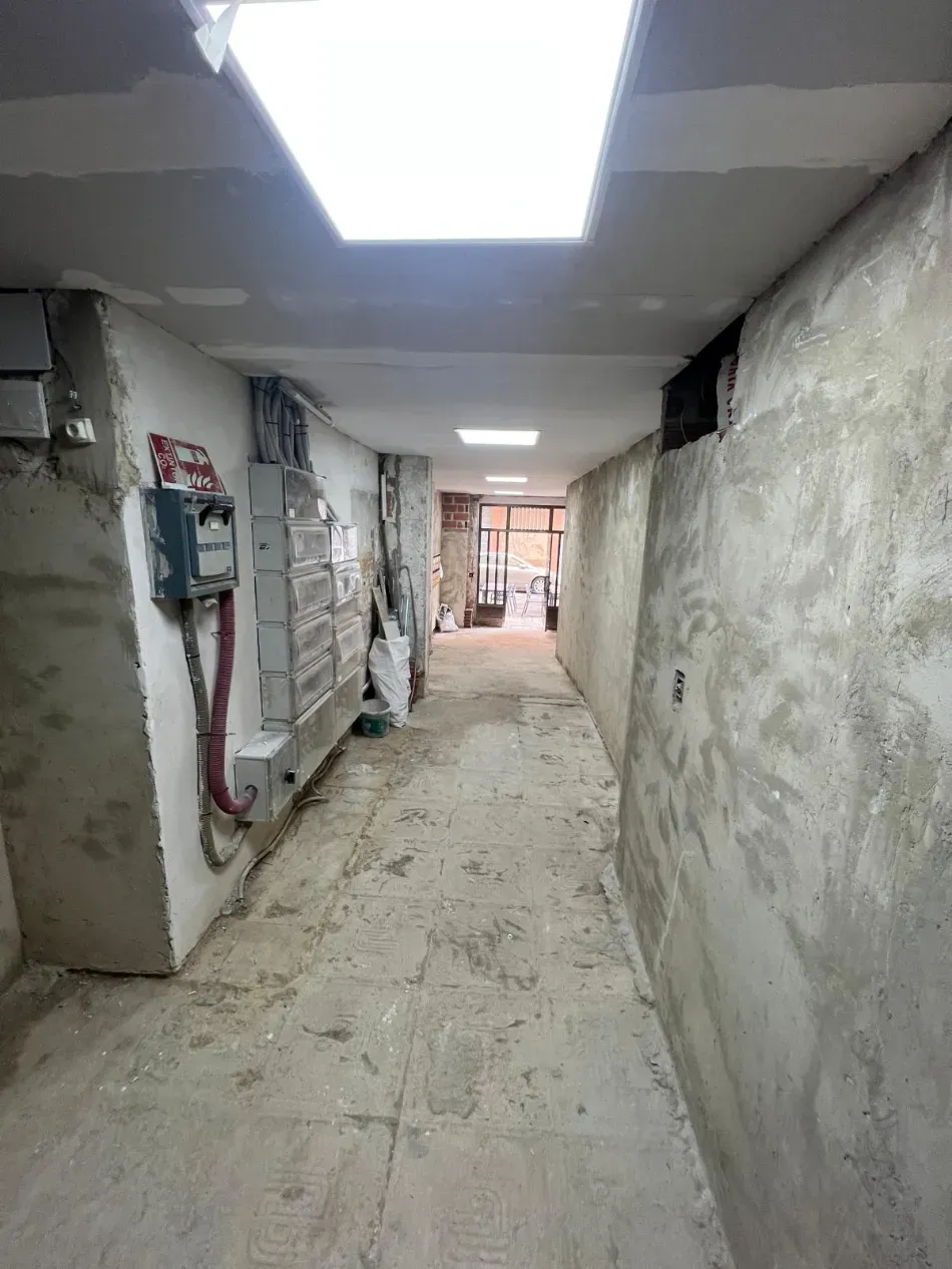 Pasillo estrecho sin terminar, con paredes desnudas, instalaciones expuestas y luces fluorescentes en el techo, que conduce a un área abierta.
