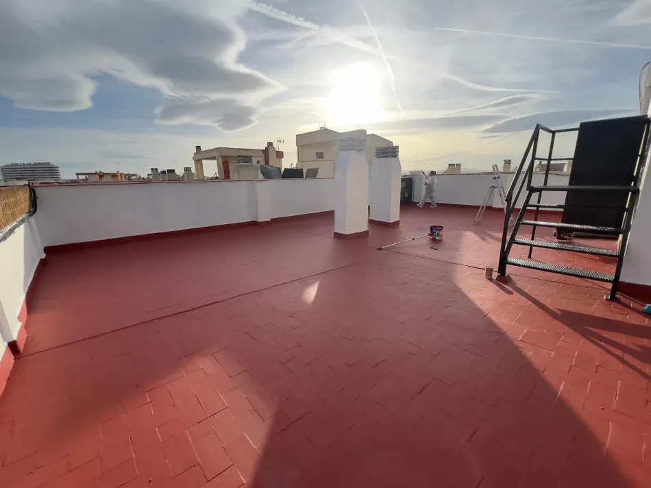 Terraza en la azotea con suelo de baldosas rojas, paredes blancas y una escalera metálica bajo un brillante cielo de puesta de sol.