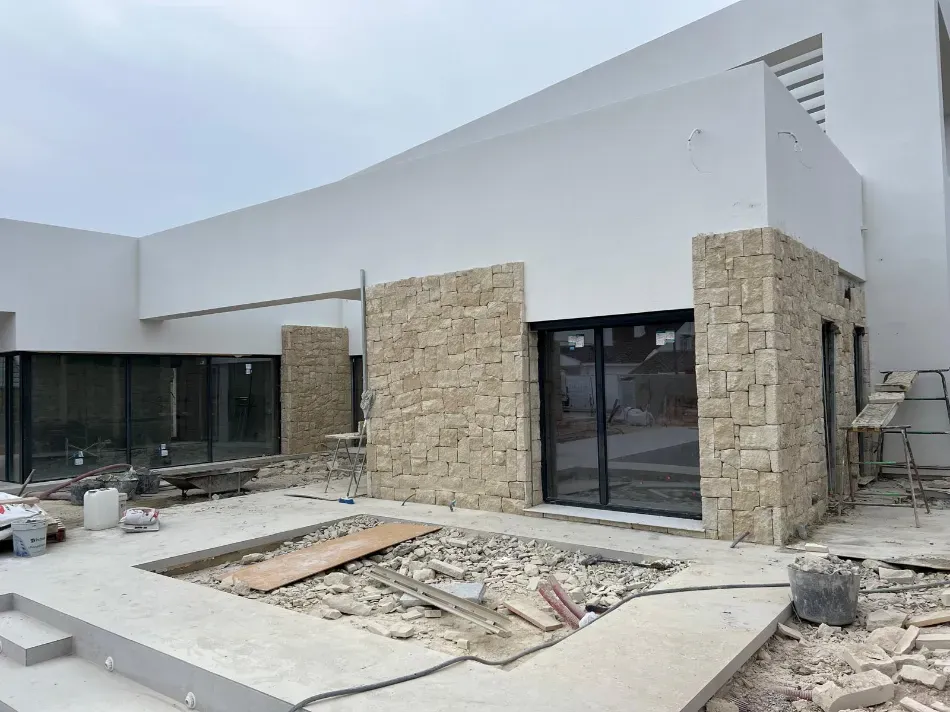 Exterior de un edificio moderno en construcción con columnas de piedra, puertas de vidrio y pasarela de hormigón sin terminar.