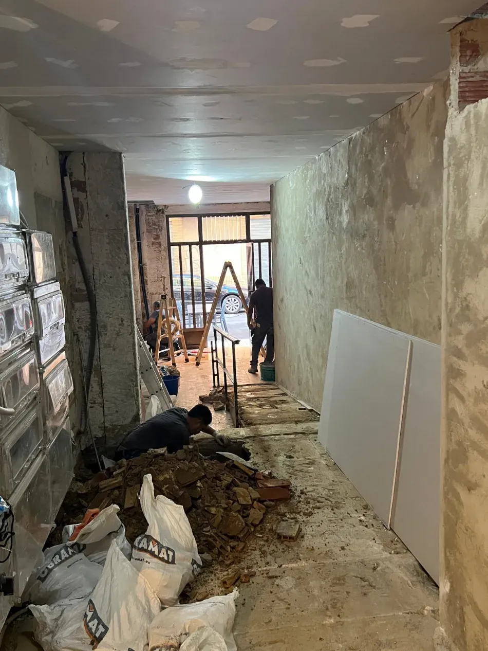 Pasillo estrecho en obras, con escombros, herramientas y trabajadores cerca del fondo.