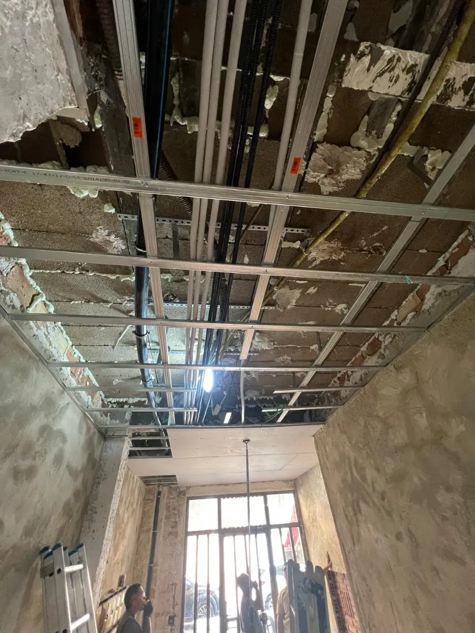 Obra en construcción en el interior, con vigas de techo a la vista, cables eléctricos colgando y trabajadores cerca de una puerta.