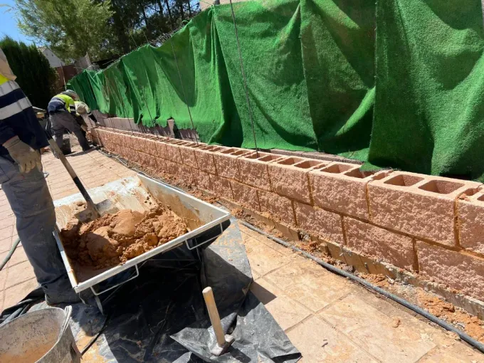 Un trabajador empuja una carretilla junto a un muro de contención de ladrillos cubierto con una lona verde durante la construcción.